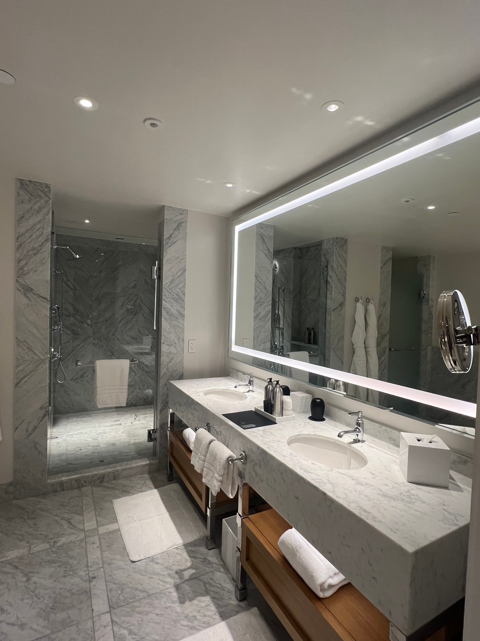 A view of a spacious, minimally designed bathroom with gray marble walls and counters, tile floors and a brightly lit mirror.