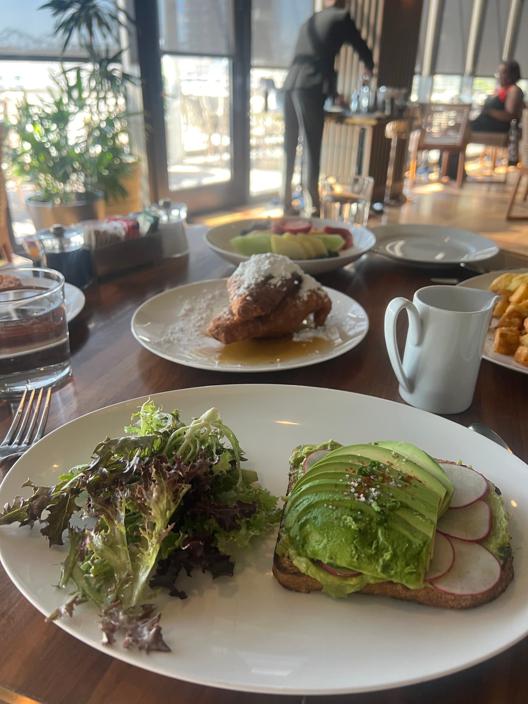 Food view of avocado toast and greens, a pastry and a coffee cup on a wooden table.