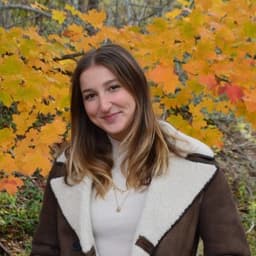 a young woman with brown hair stands in front of a yellow-leaf tree