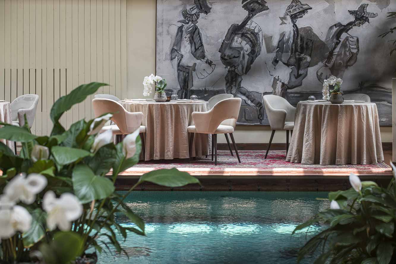 View through plants of a swimming pool with round dining tables set up to one side with tablecloths and a large piece of artwork on the wall behind.