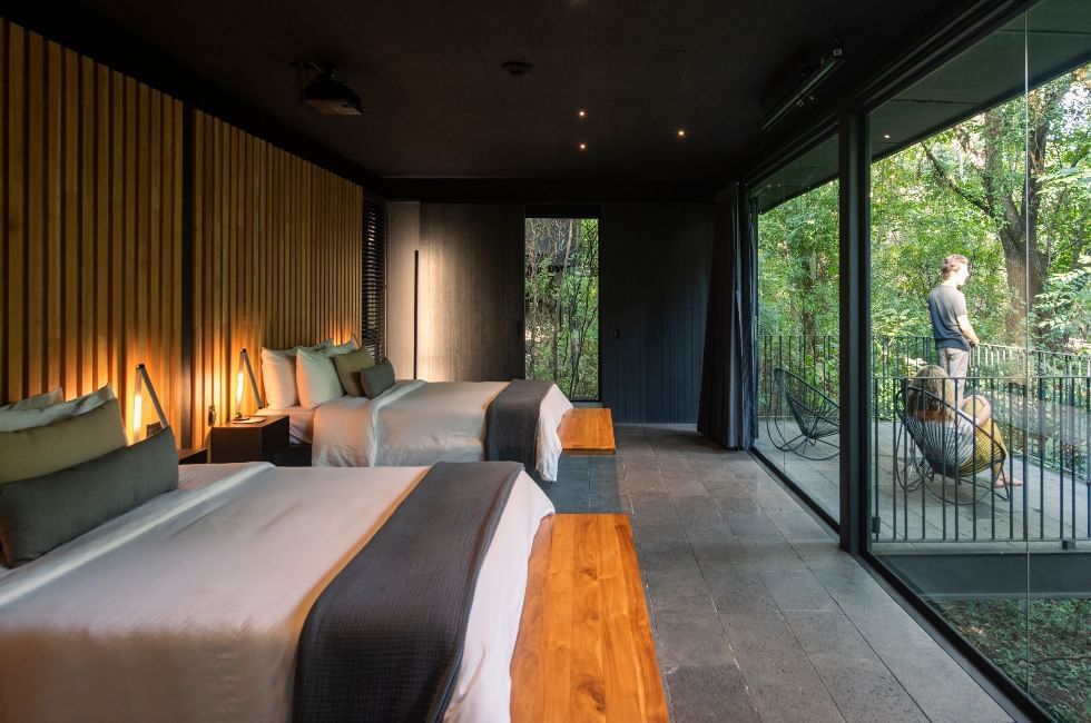A cozy hotel room in tones o gray with floor-to-ceiling glass windows and a terrace and jungle views.