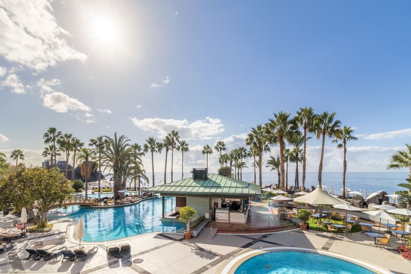 A view of the pool area at the Royal Savoy with tall trees and a kiosk with a green roof between the pools.