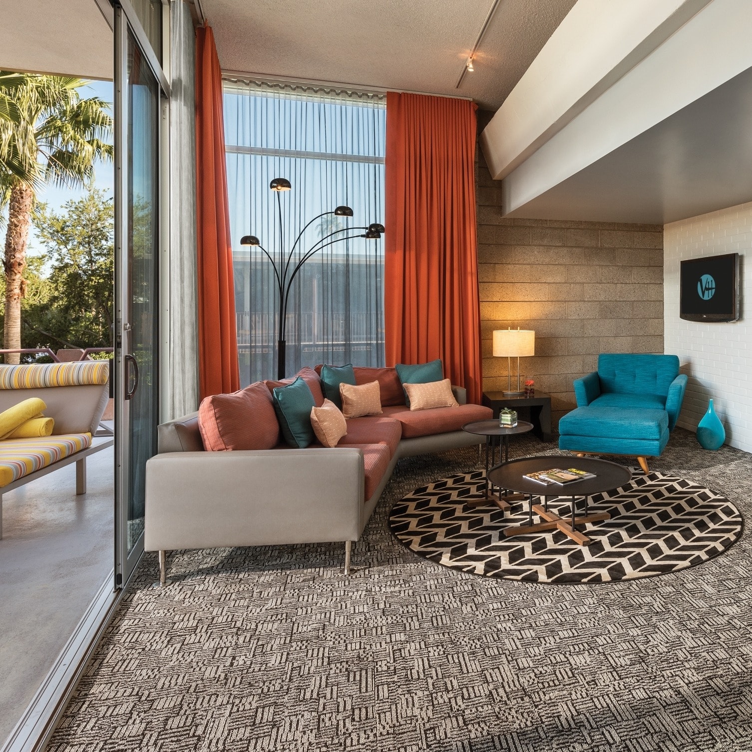 A suite at the Hotel Valley Ho, with a couch and blue chair in the living room, orange curtains, brick walls and a terrace with a palm tree in the distance.
