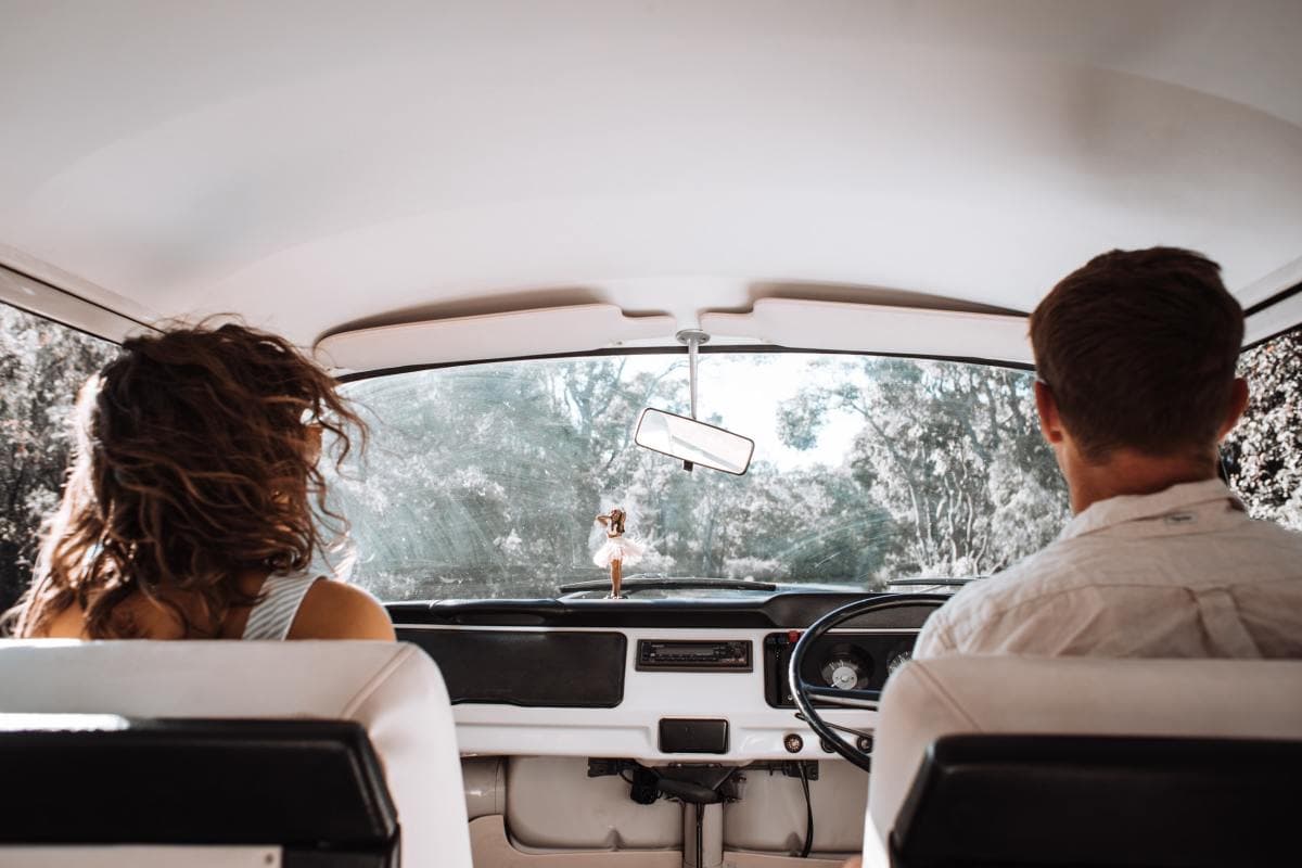 Woman and man sitting in white car surrounded by green trees with hula girl on the dashboard