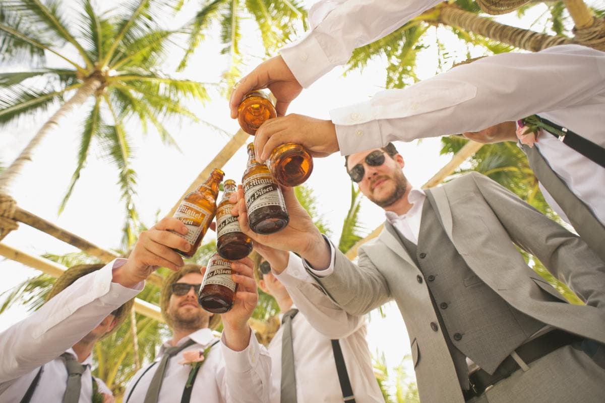 Five men wearing suits and sunglasses cheers with beers while standing beneath palm trees