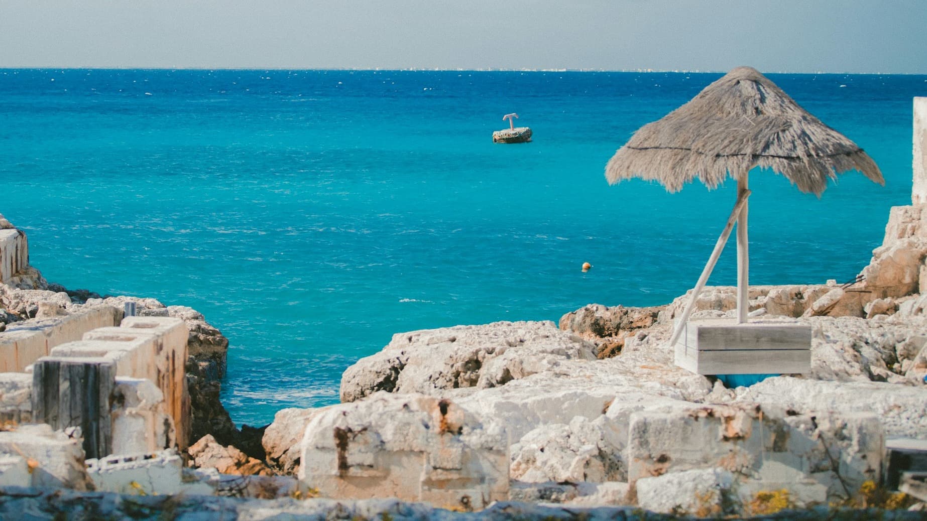 A rocky shore with a straw umbrella and beautiful turquoise ocean with a boat in the distance