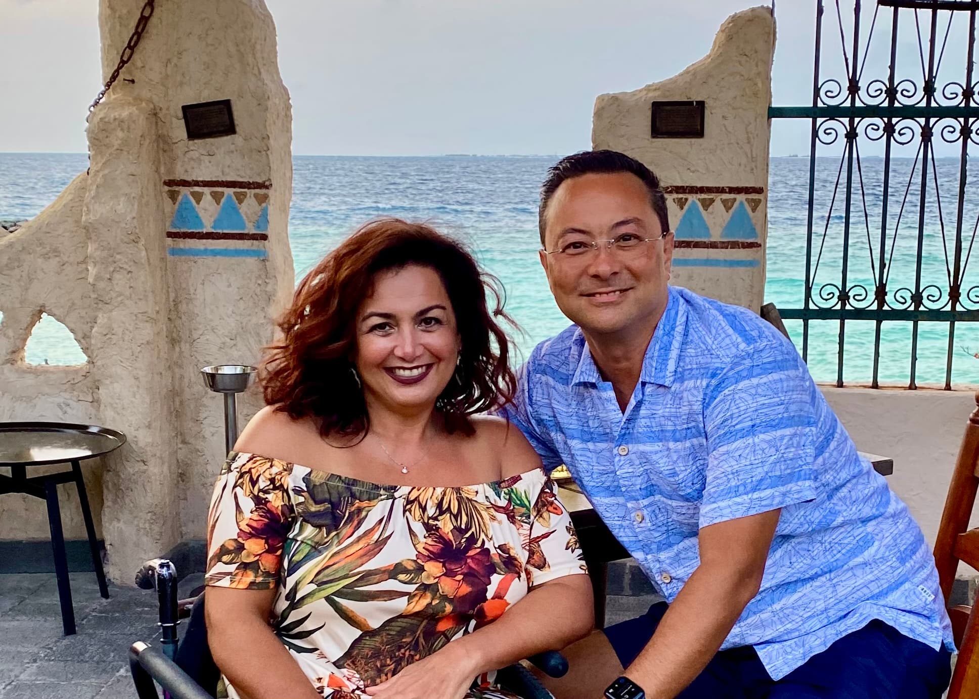 A couple, Glenn and Judy Tudor, sit next to each other smiling with a beach in the background