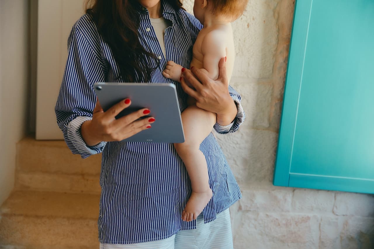 a woman in a striped shirt holds a baby in one hand an iPad in another