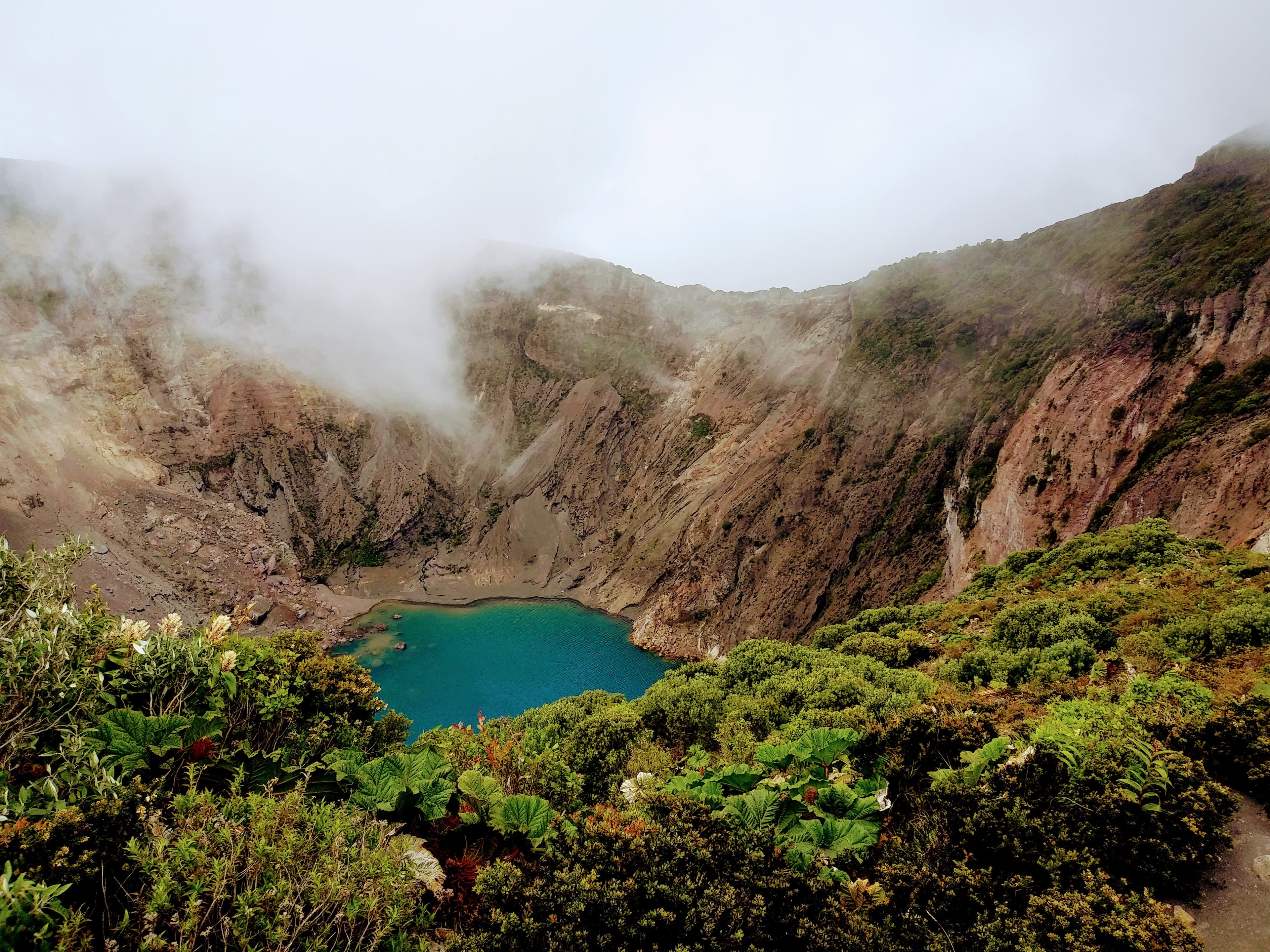Brown crater with white wispy clouds and a blue lake with green and yellow shrubs