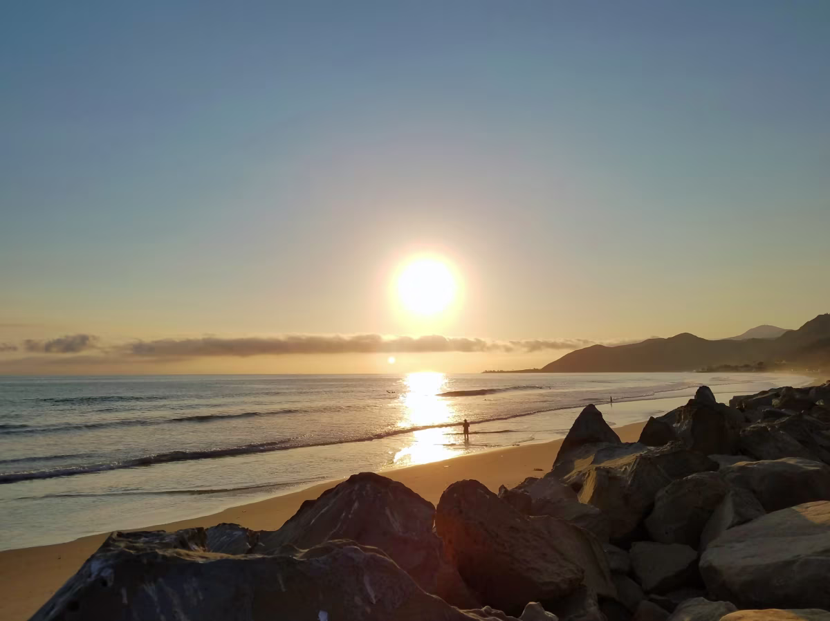 A person walking along a sandy beach rocky coastline next to it as the sun begins to set.