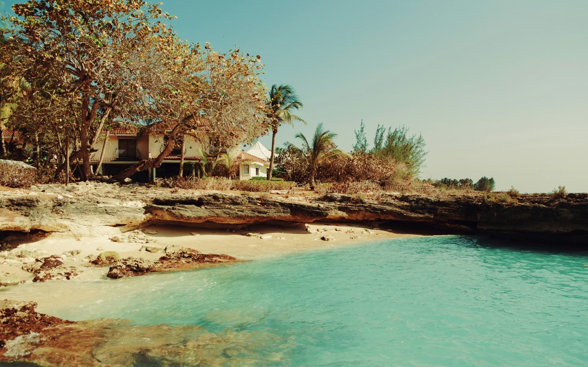 A view of the beach with bright teal water, white sand, palm trees and a house on the shore.