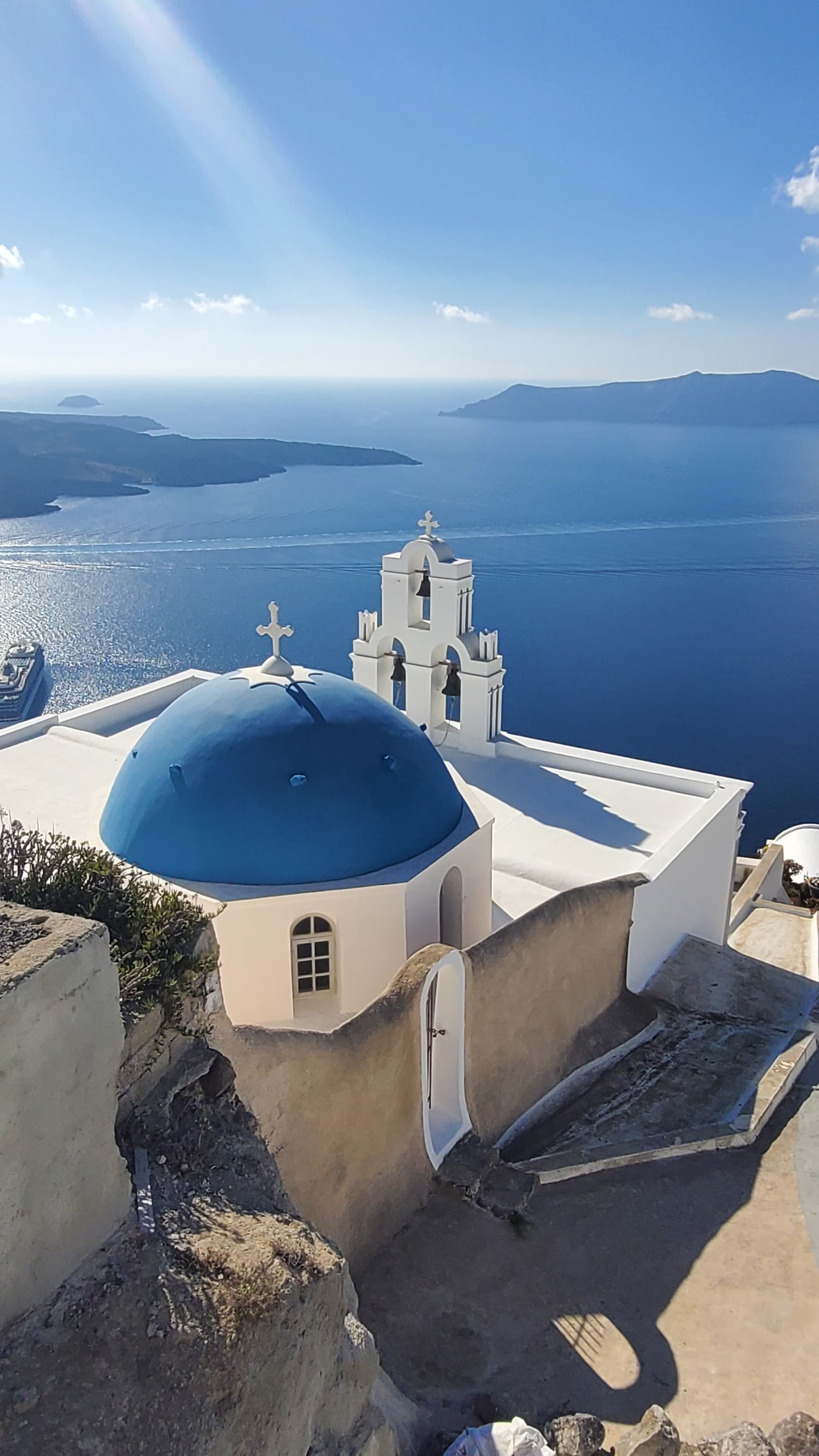 A view of the coastline of Santorini, Greece, with bright white buildings and the famous blue dome overlooking the ocean on a sunny day.