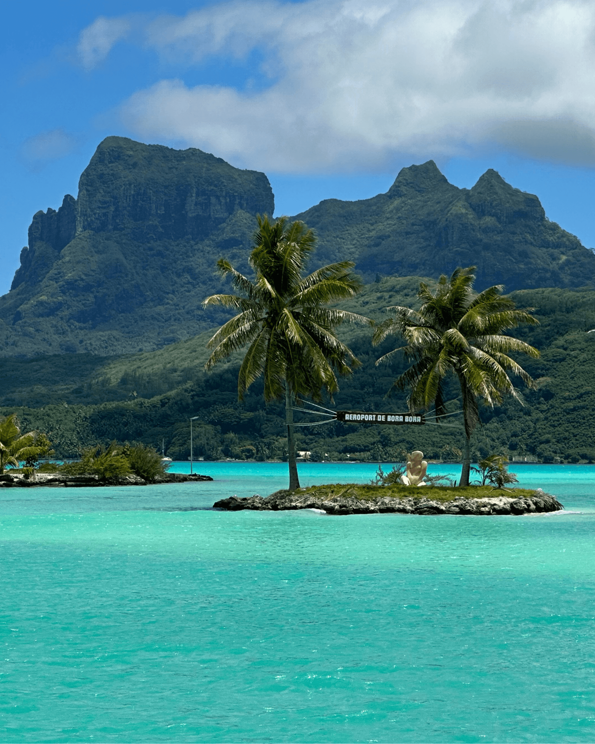 A view of a bright teal blue ocean with a small island with two palm trees and a large mountain in the distance on a partly cloudy day.