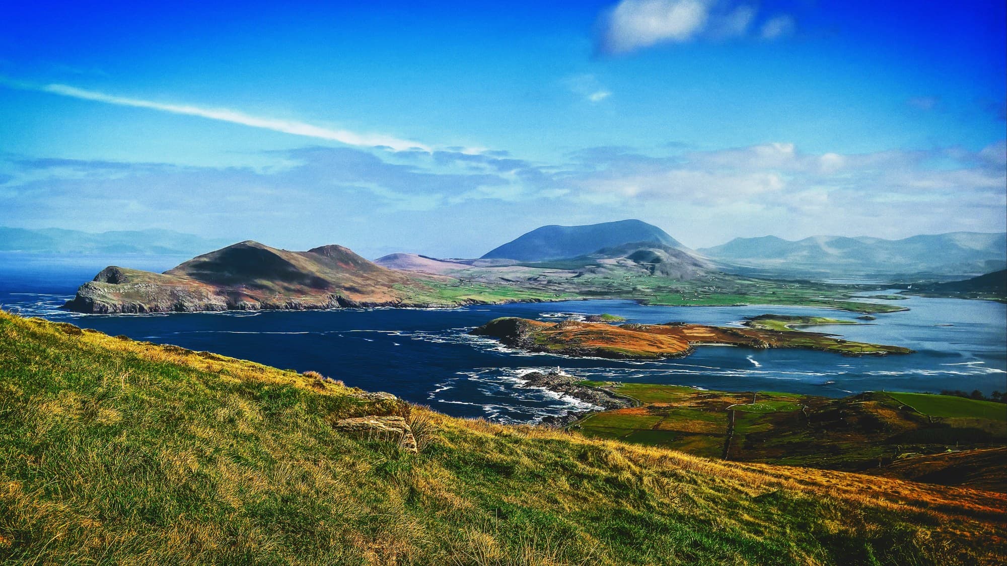 A view of a grassy hillside with the ocean and large mountains and a big blue sky in the distance. Skellig Ring on Geokaun Mountain on Valentia Island, Foilnageragh, Co. Kerry, Ireland