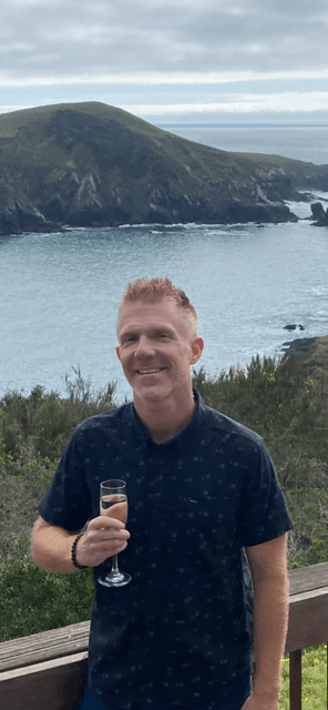 A person standing by a railing smiling for a photograph with a view of the water and a mountain in the distance