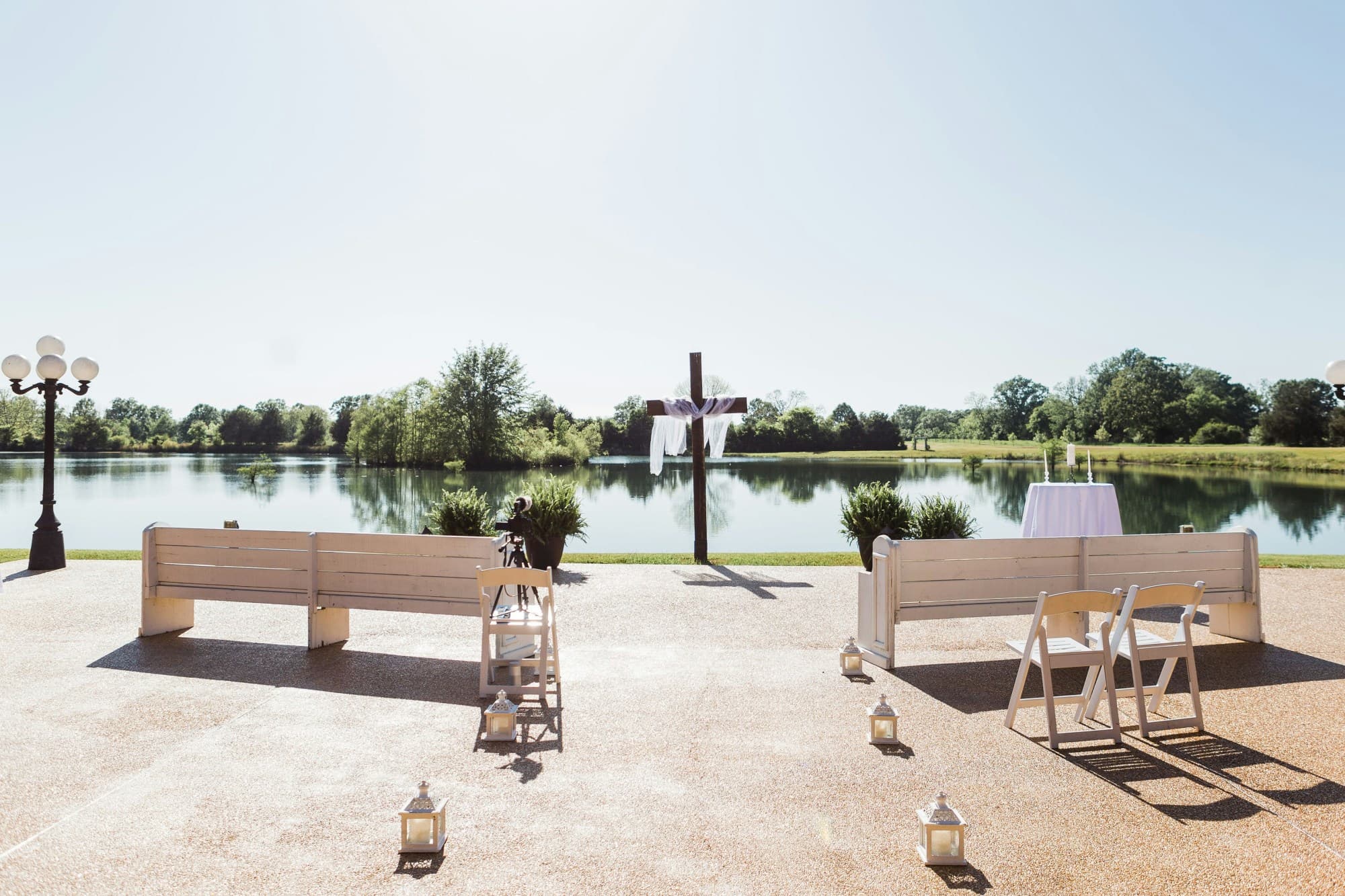 A wedding ceremony staged with two pews on either side of an aisle lined with candles and large cross at the altar, there is a lake in the background on a sunny day.