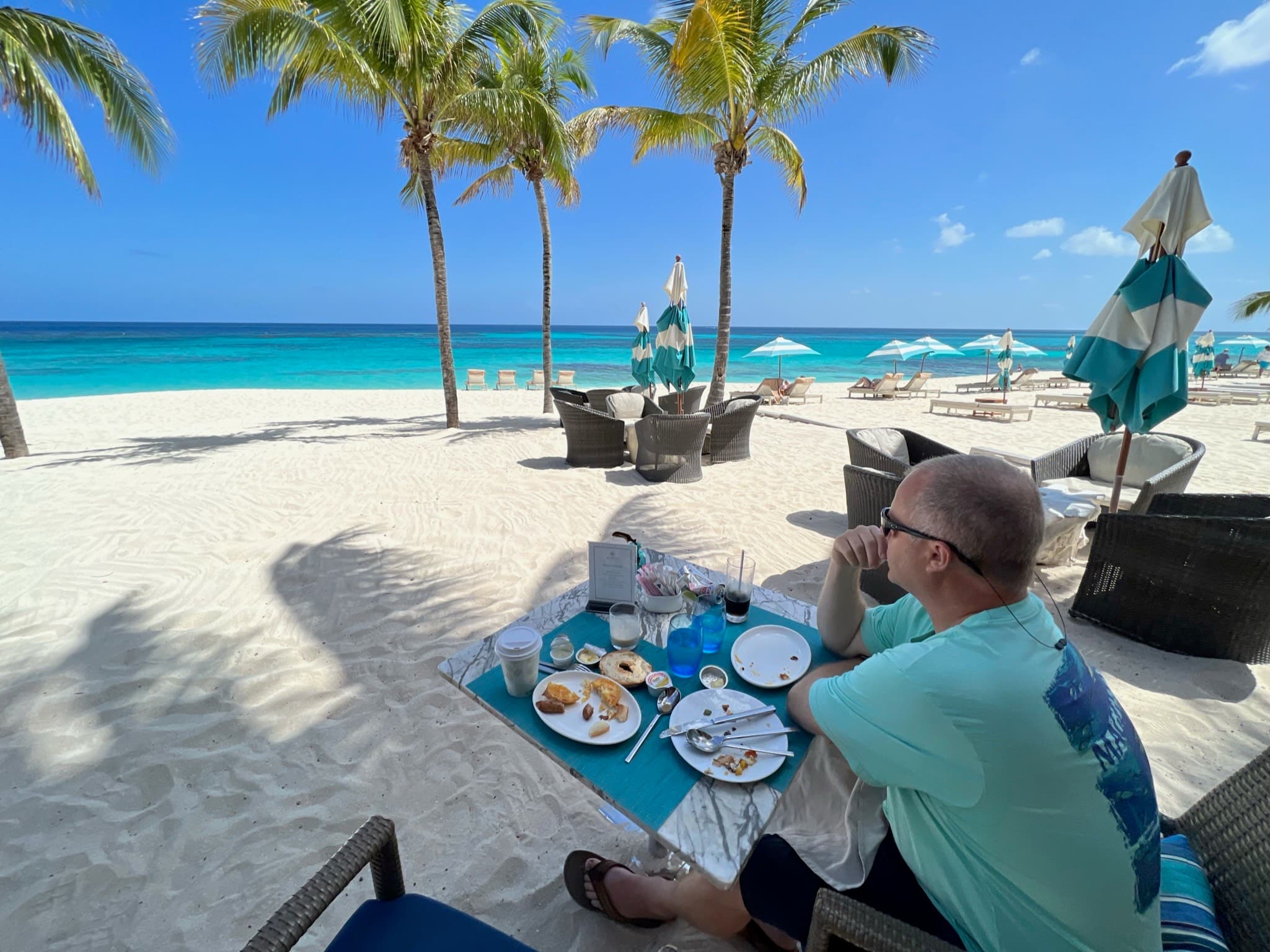 A man sits next to a low table with food on a beach and looks out at the ocean.