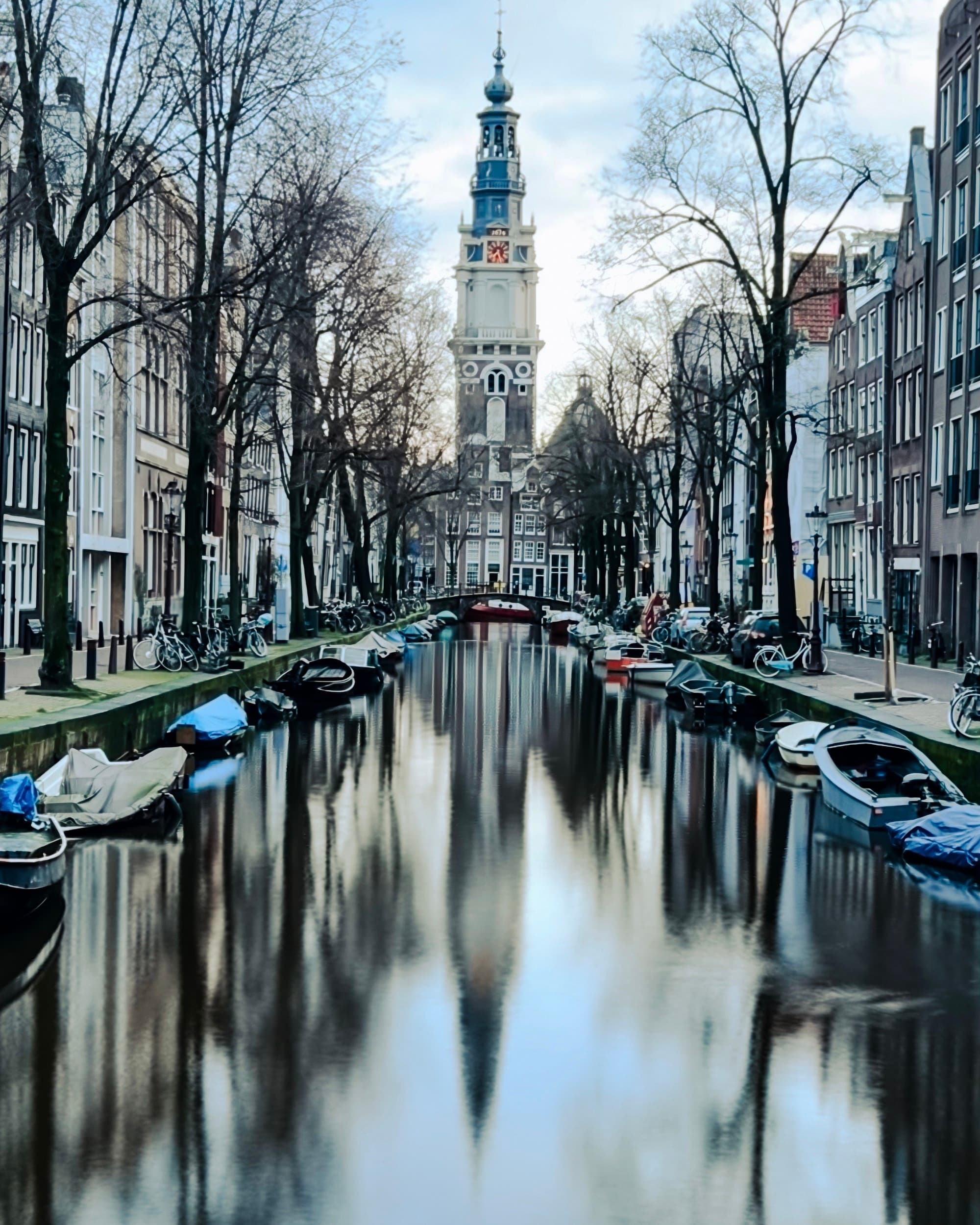 A canal in Amsterdam gleams in the sunlight as trees reflect on the water with boats lining the shore.