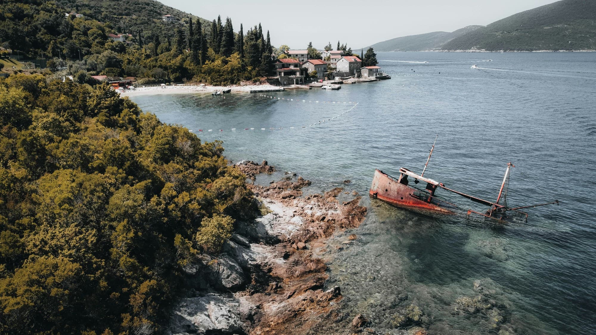 Shipwreck Beach in Rose at the entrance to the Bay of Kotor, with a red boat partially submerged in water off of a tree-covered hillside.