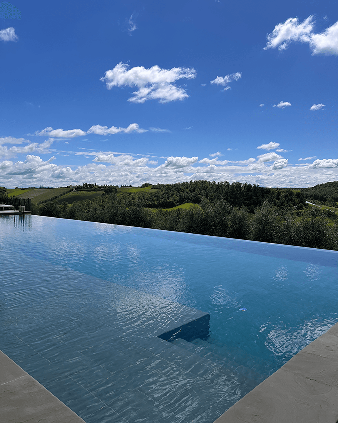 A point of view shot of an infinity pool overlooking the rolling, green hills of Tuscany on a sunny day.