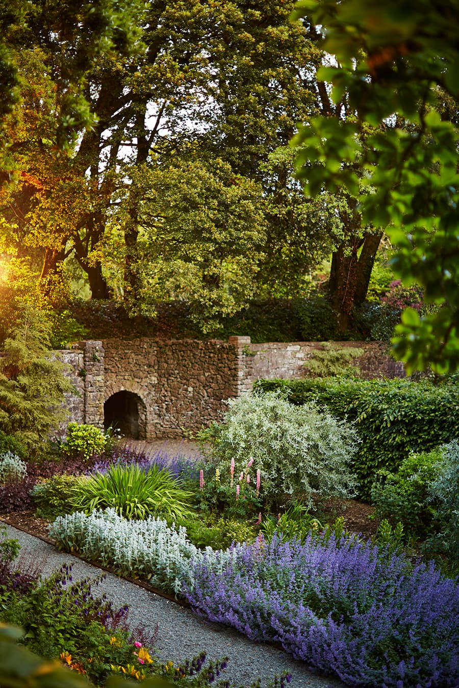 A stone wall with an archway surrounded by a lush shade garden with greenery and purple flowers at the Ashford Castle, one of the best 5 star hotels Ireland.