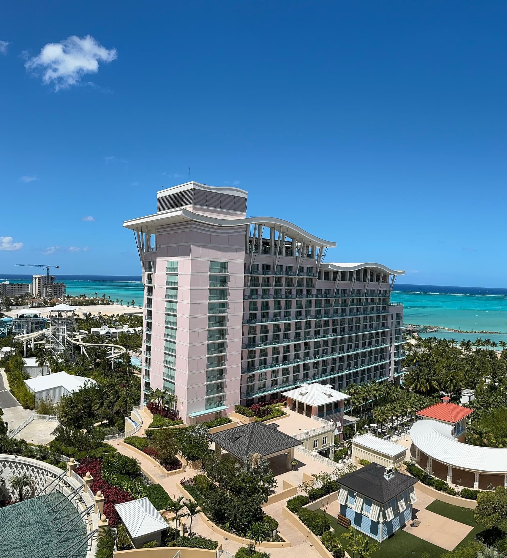 An image of a tall resort building surrounded by smaller buildings, paths and trees with a turquoise blue body of water in the background.