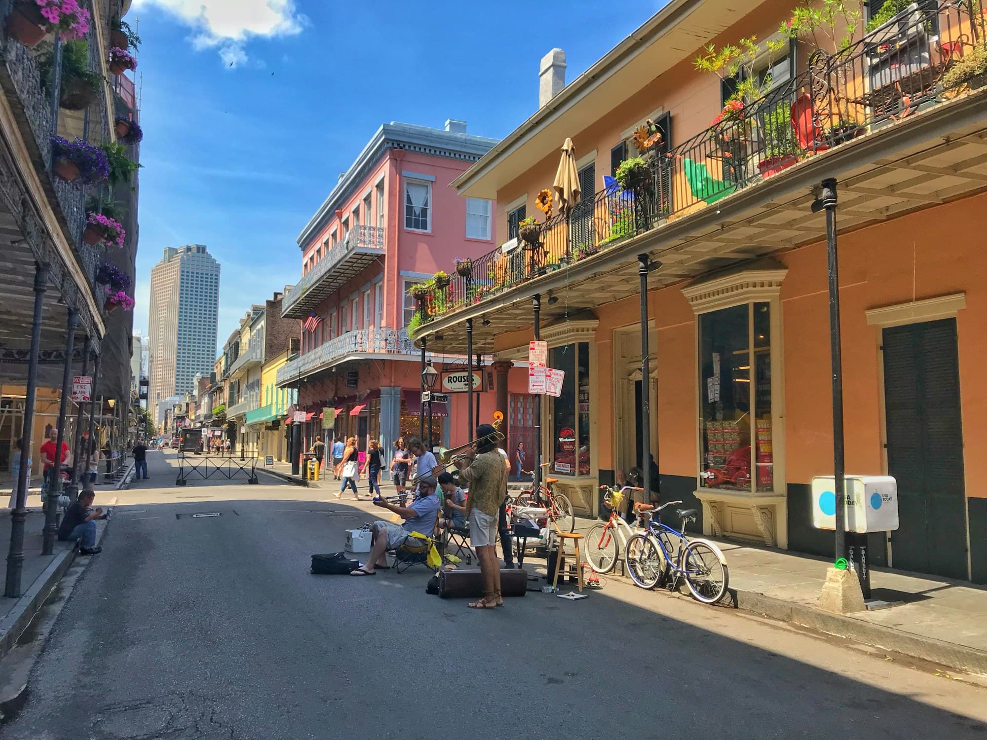 A view of a vibrant street full of people in New Orleans. There are also bicycles and orange and pink buildings with metal balconies, decorations and plants in the surrounding areas.