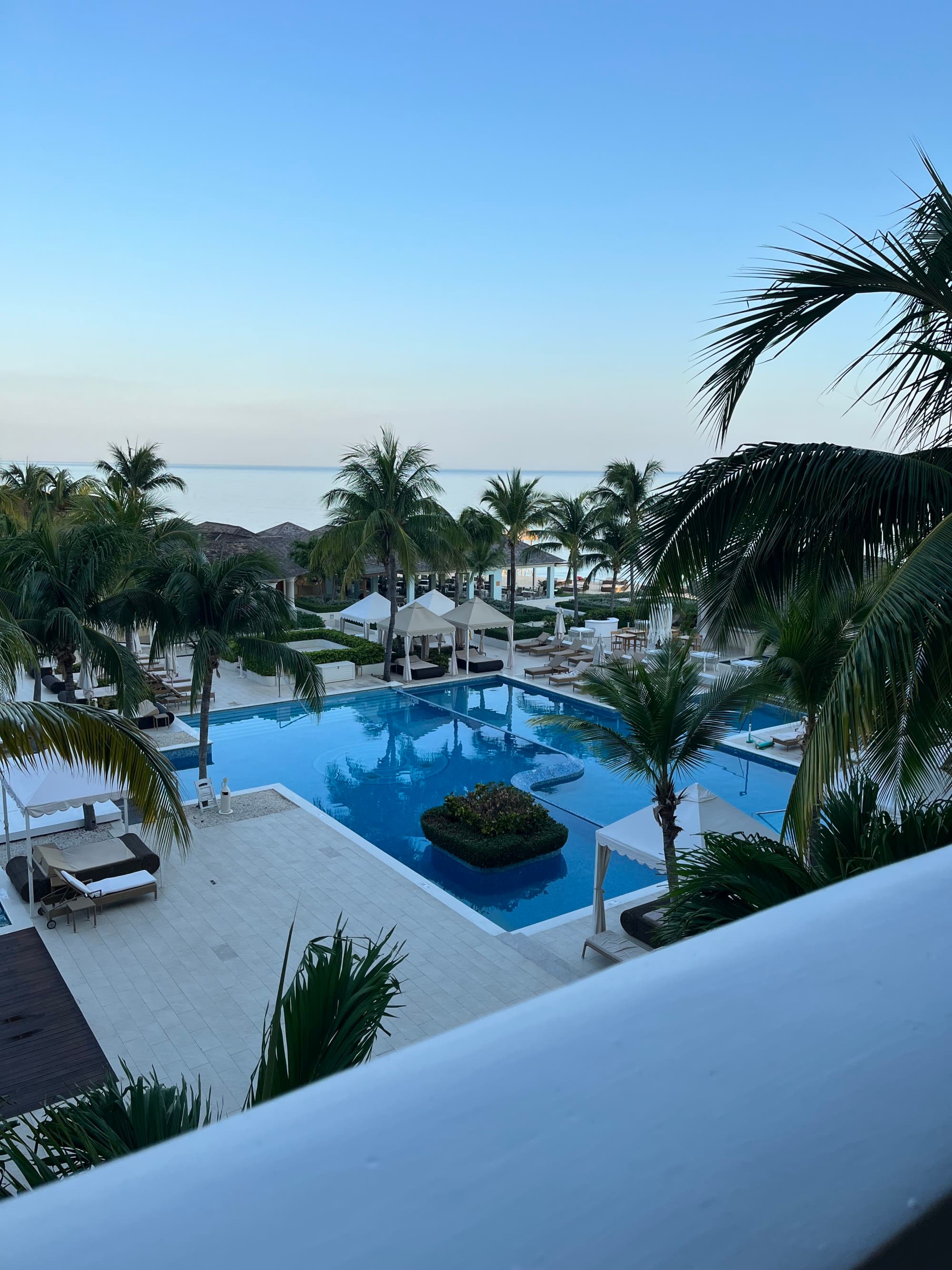 A picture of a pool with palm trees, lounge chairs and cabanas in the surrounding area.