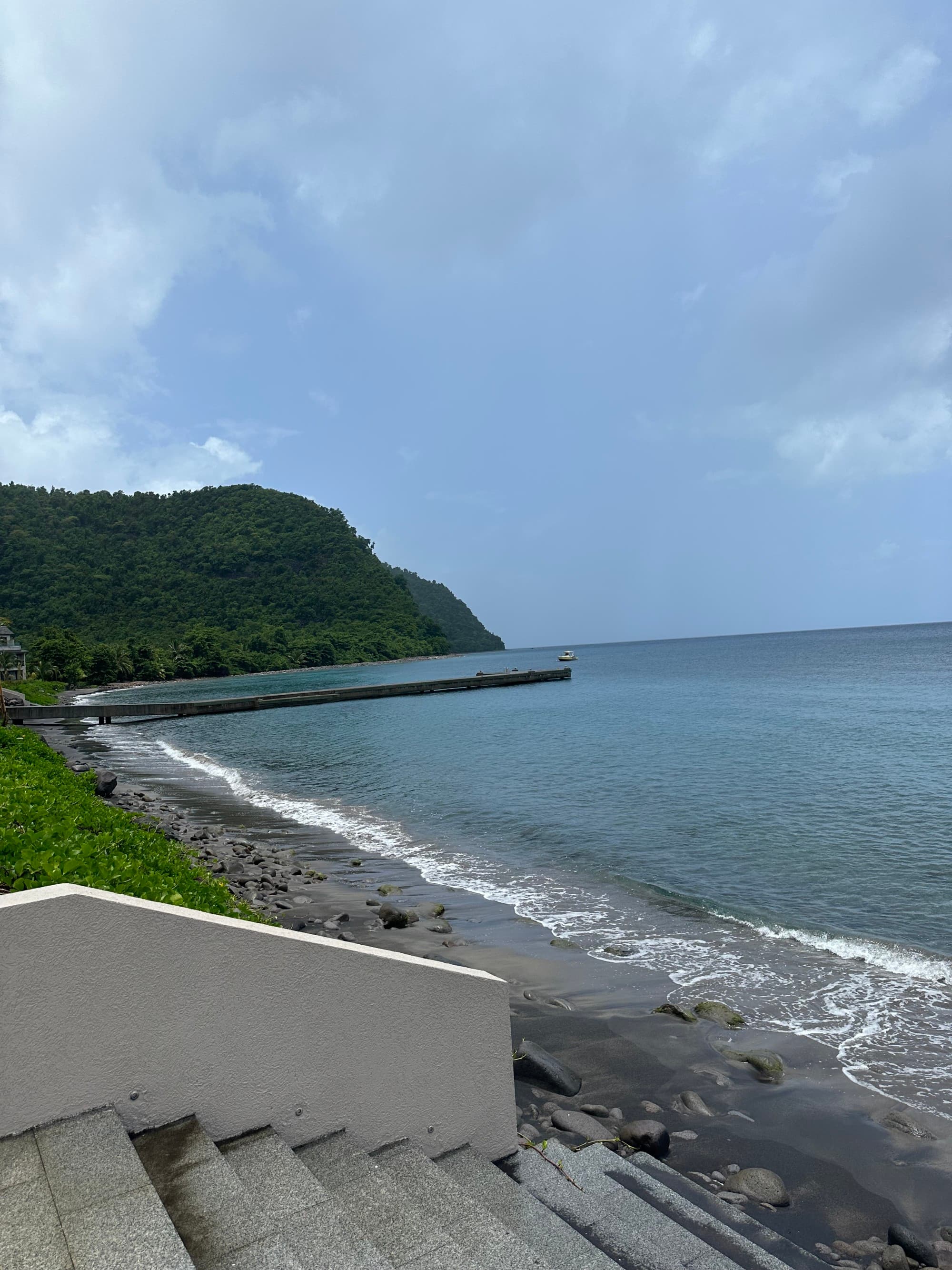 A view of white stairs leading to a dark sandy beach and blue sea. There is a green mountain in the background.
