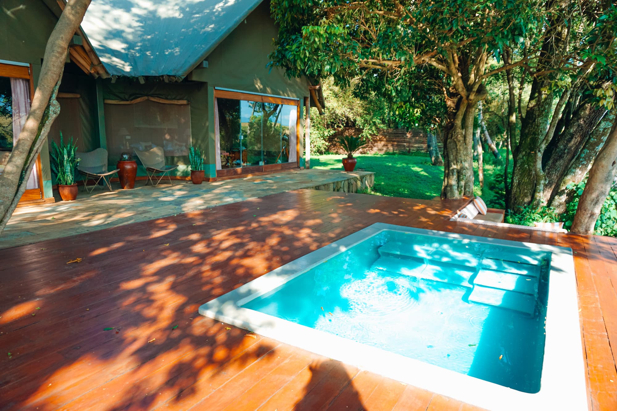A patio with a pool, trees, wood fooring and a house with potted plants around it on the left side of the image.