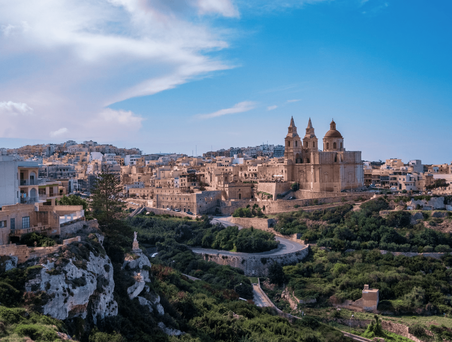 A beautiful aerial view of stone buildings tucked into a coastal array of rocks, trees and winding roads in Malta.