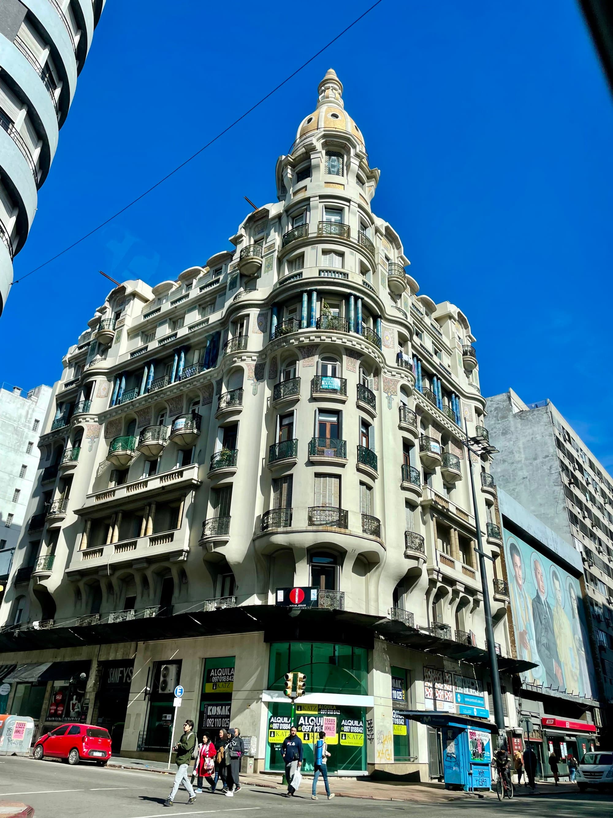 A bottom-up view of a white building exterior with a blue sky behind it. There are two people walking in front of it.