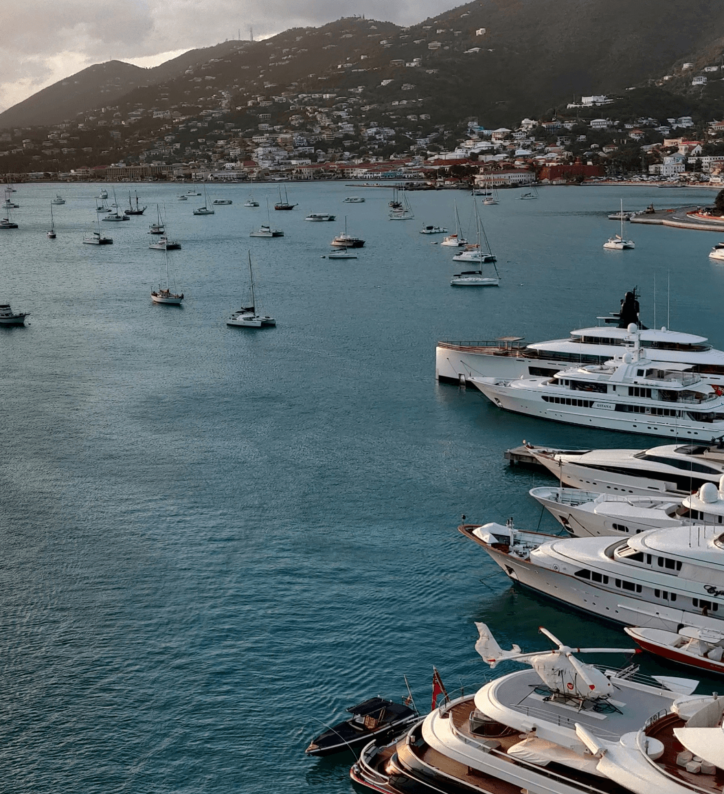 A view of yachts anchored in a harbor with sailboats and buildings tucked into a mountain in the background.