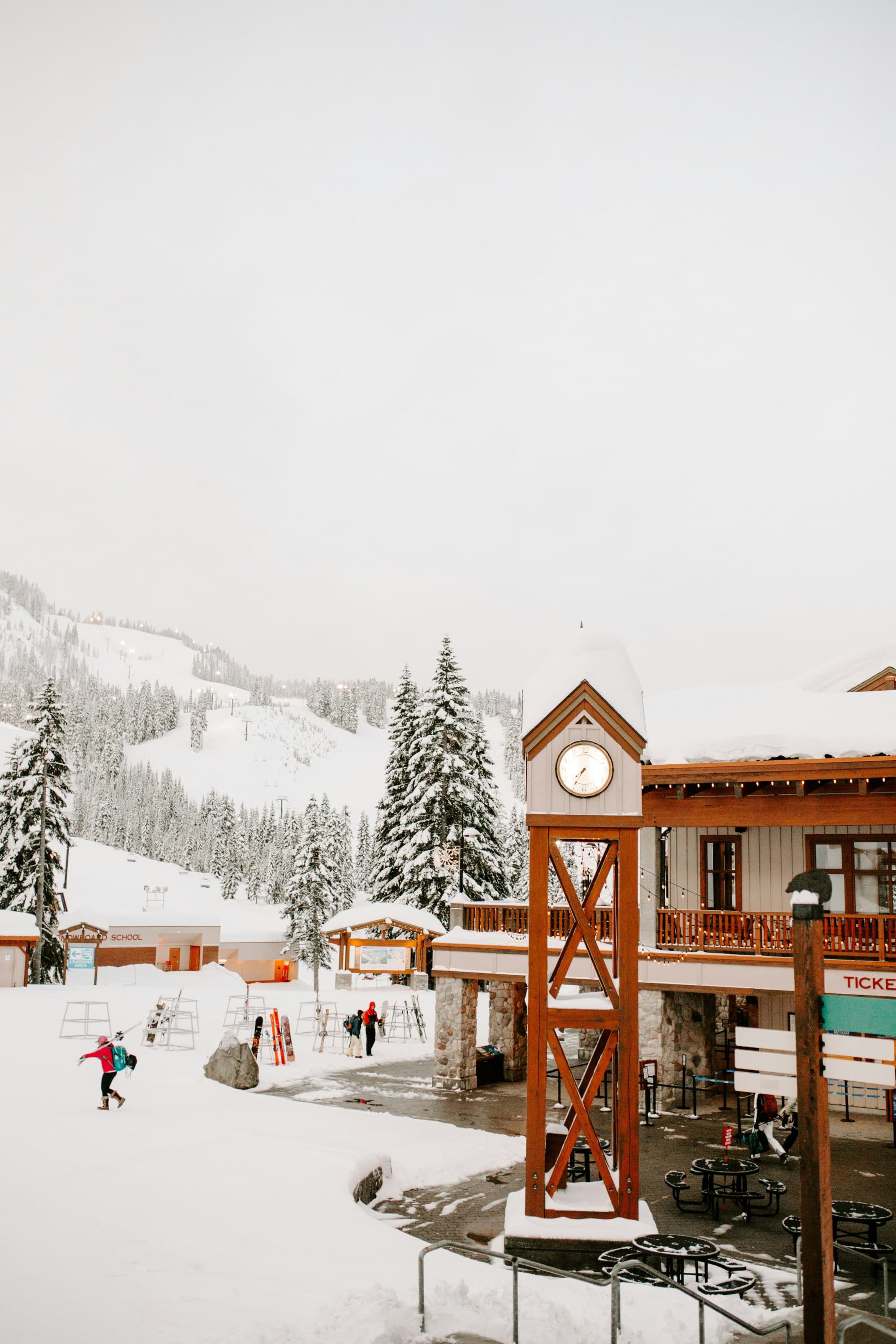A view of a ski lodge, clock tower and people walking on the snowy terrain in the distance towards snowy pine trees and a ski hill that escapes into the fog.