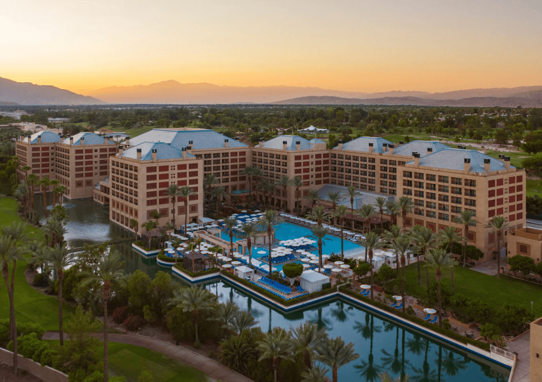An aerial view of a resort surrounded by palm trees, a pool with lounge chairs and umbrellas, a bar a body of water and mountains beneath a golden sunset in the distance.