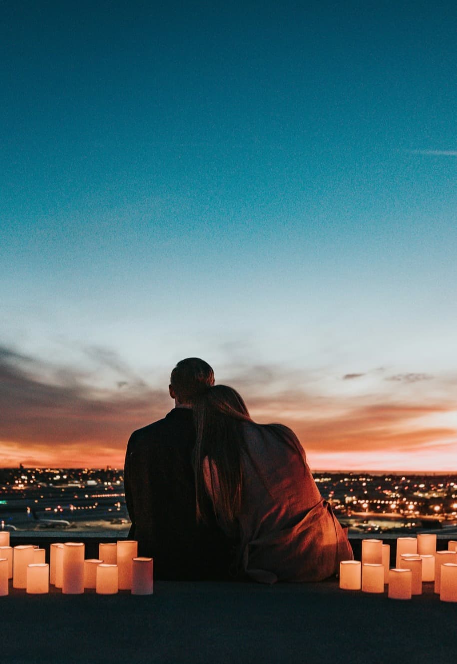 A silhouette of a couple posing while watching the sunset with candles lit around them and a city view in the distance.