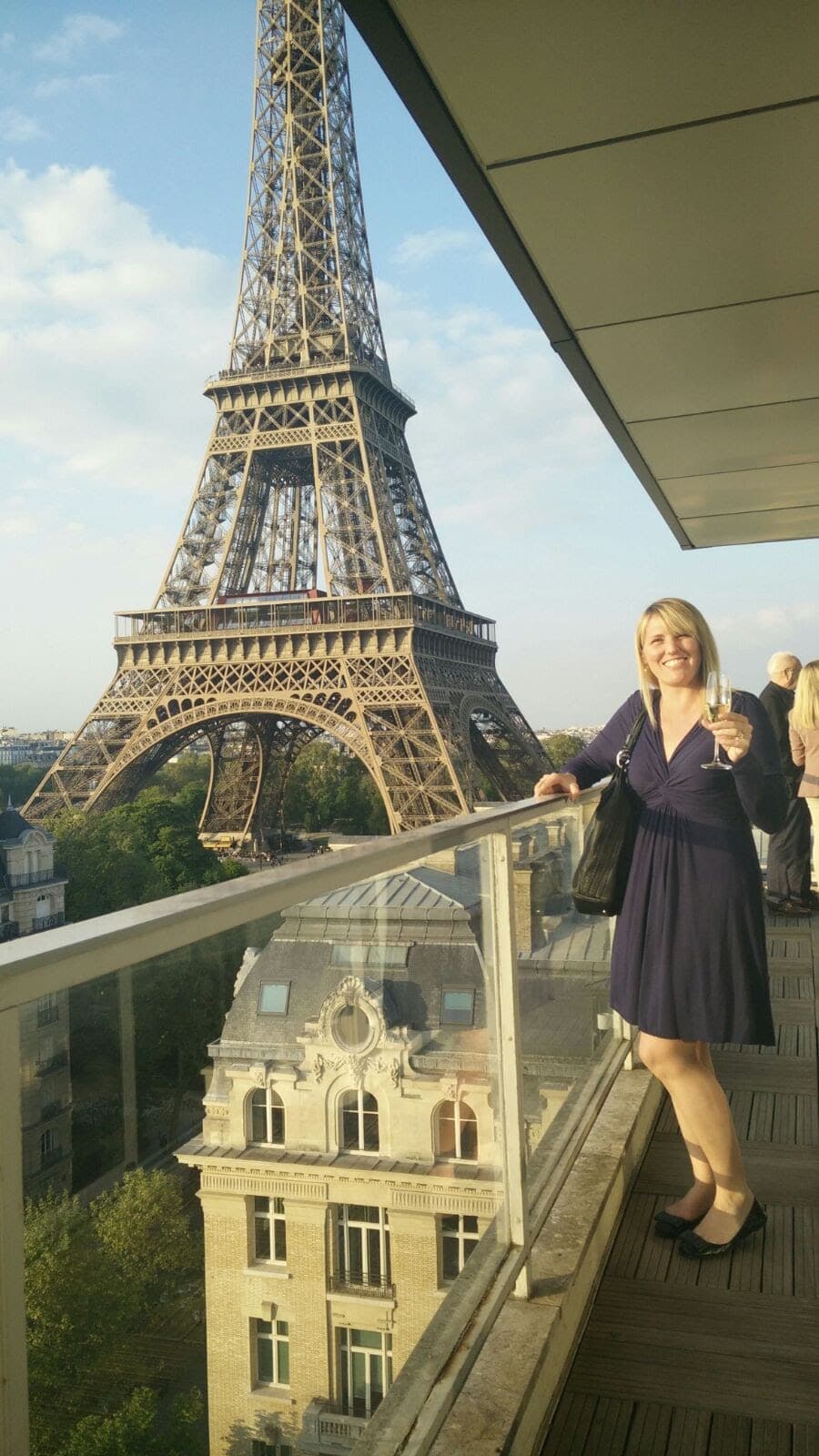 Travel advisor Heather in a blue dress standing on a balcony with a glass of champagne and the Eiffel Tower in the background in one of the best areas in Paris.