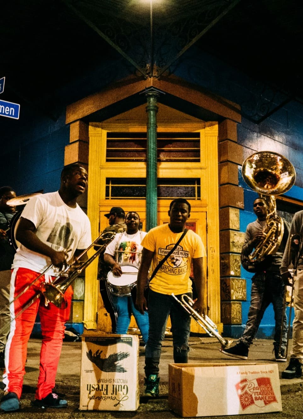 A group of people playing instruments outside in front of a yellow and blue building at nighttime.