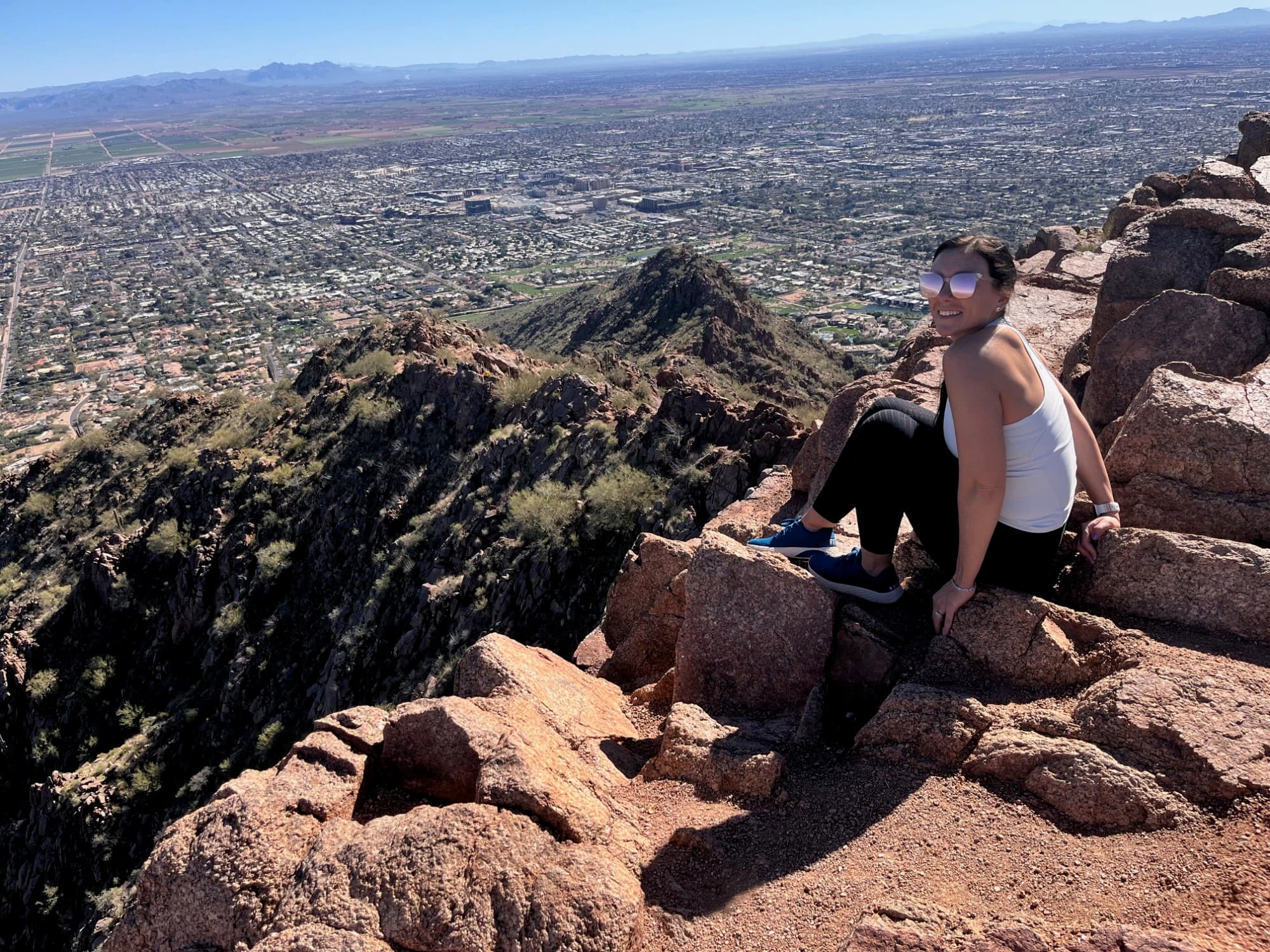Travel advisor Kelly wearing a white top and sunglasses sitting on the edge of a high rocky cliff overlooking a beautiful view of a city