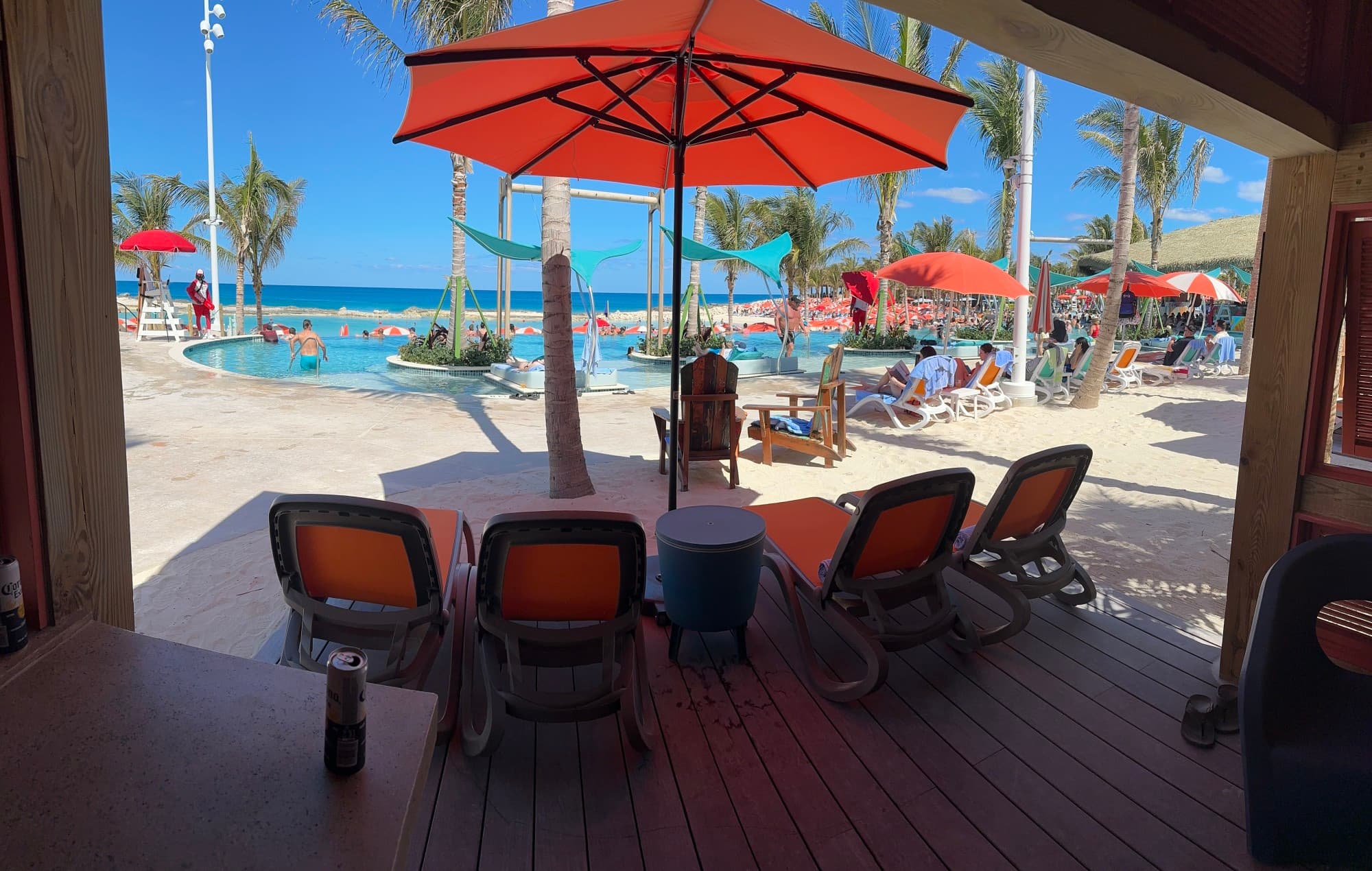 A pool with beach chairs, orange umbrellas and beachgoers in front of the ocean.