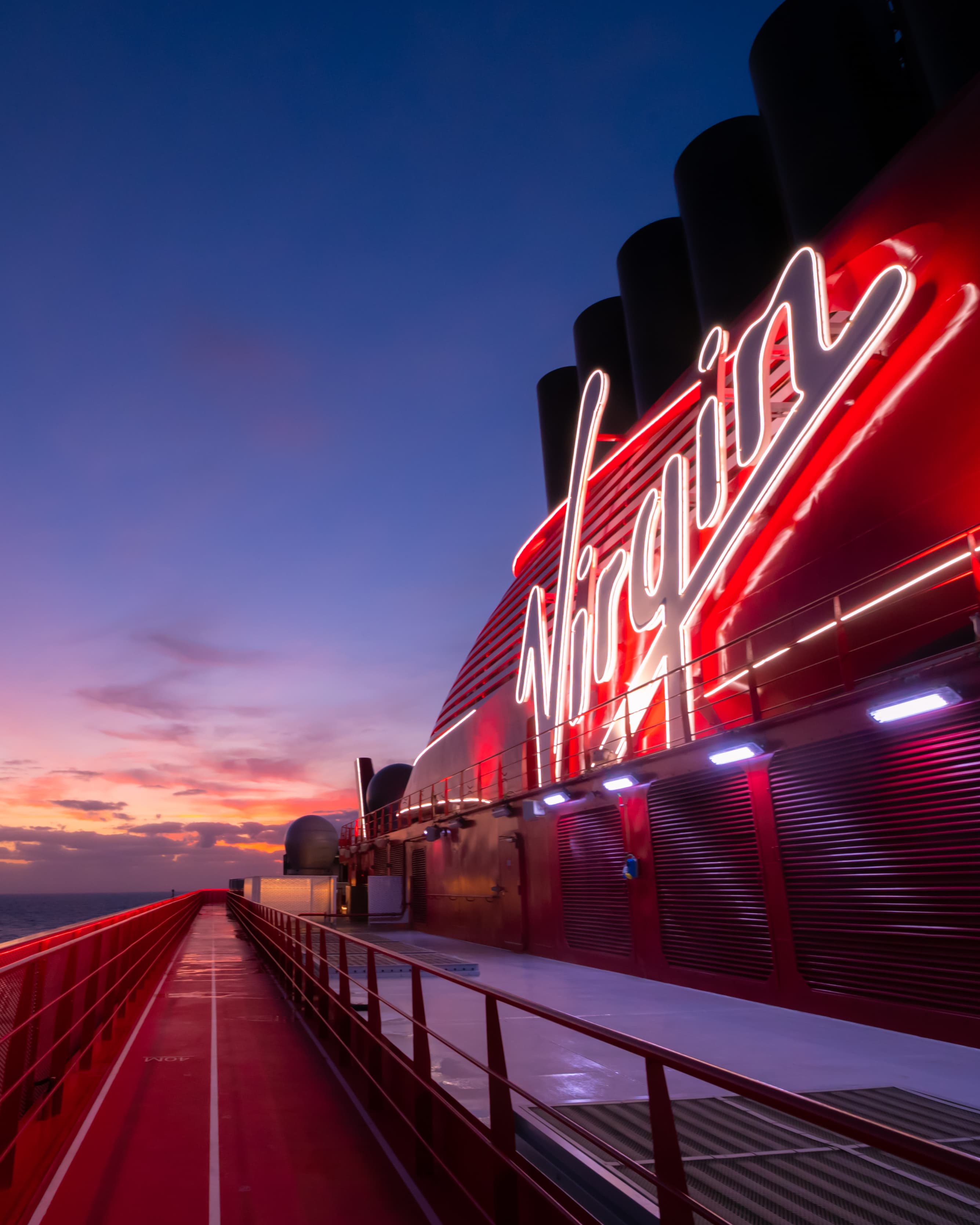 A view of a large neon red sign that says "Virgin" over the deck of a ship. There is a pink, blue and orange sunset in the distance.