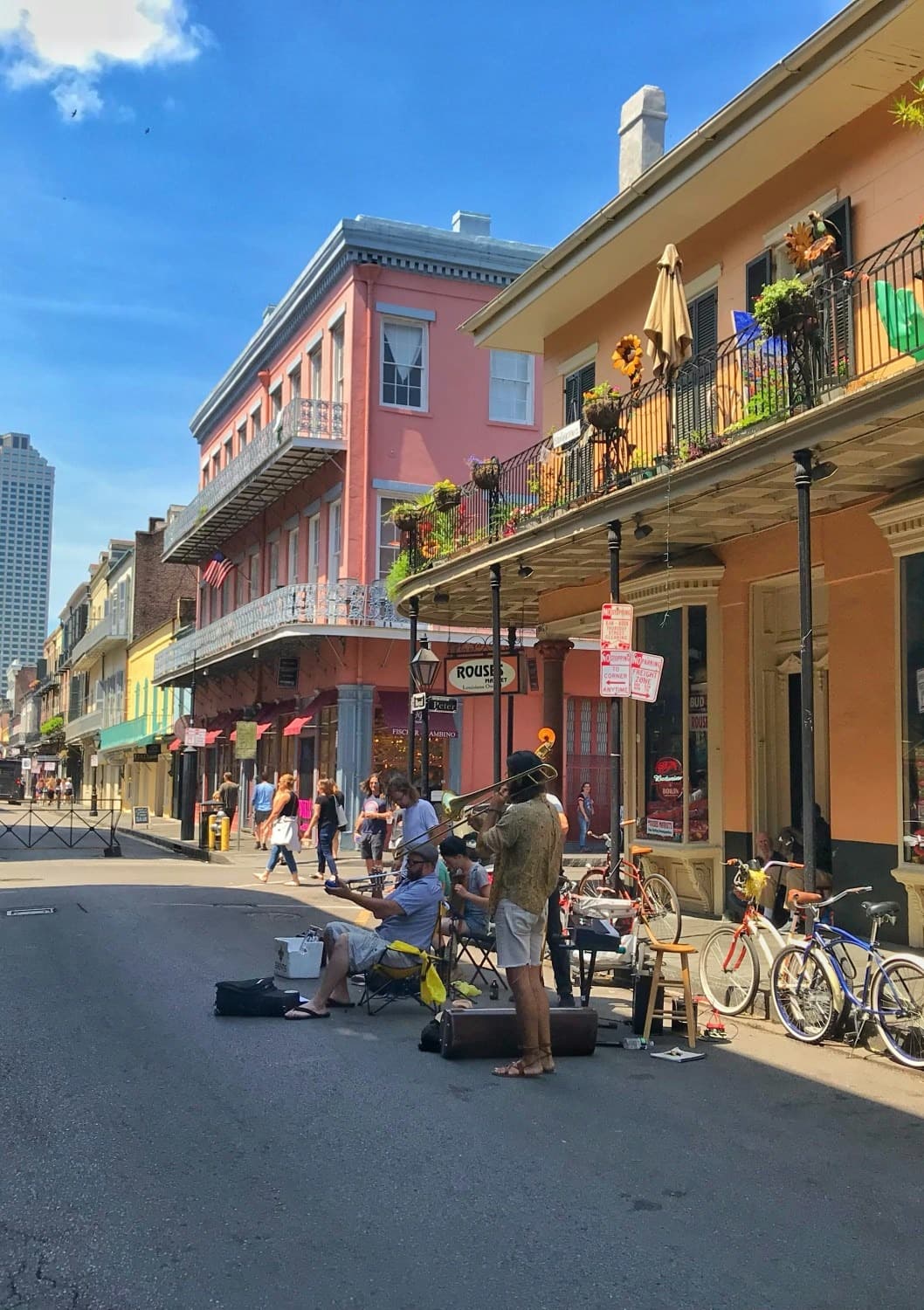 A group of people playing musical instruments on the road next to yellow and pink buildings with flags, metal balconies and plants.