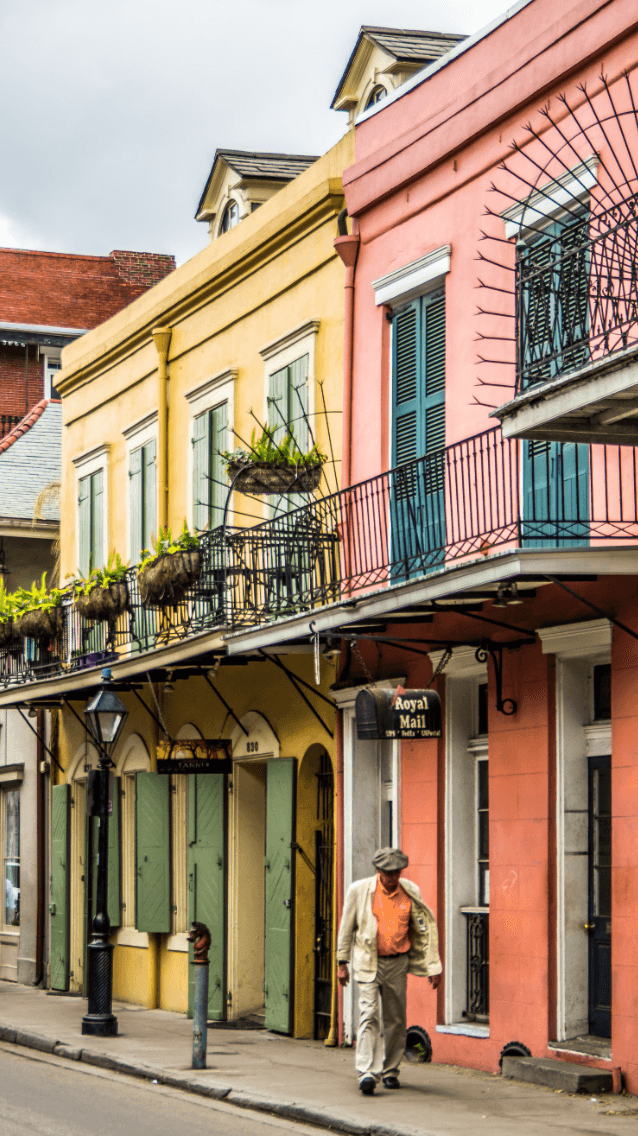 A man walking down the street next to pink and yellow buildings with shutters and metal balconies.