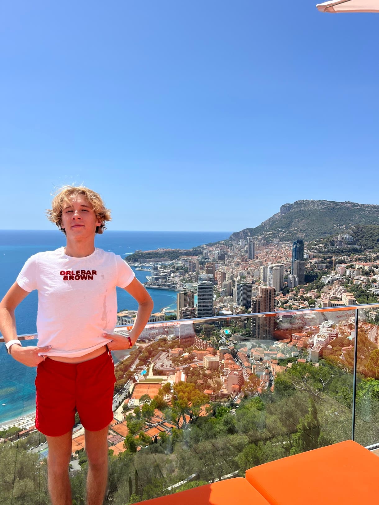 Jack wearing a white graphic tee and red shorts posing in front of a glass ledge that looks out to a view of a coastal city, mountain and blue sea.