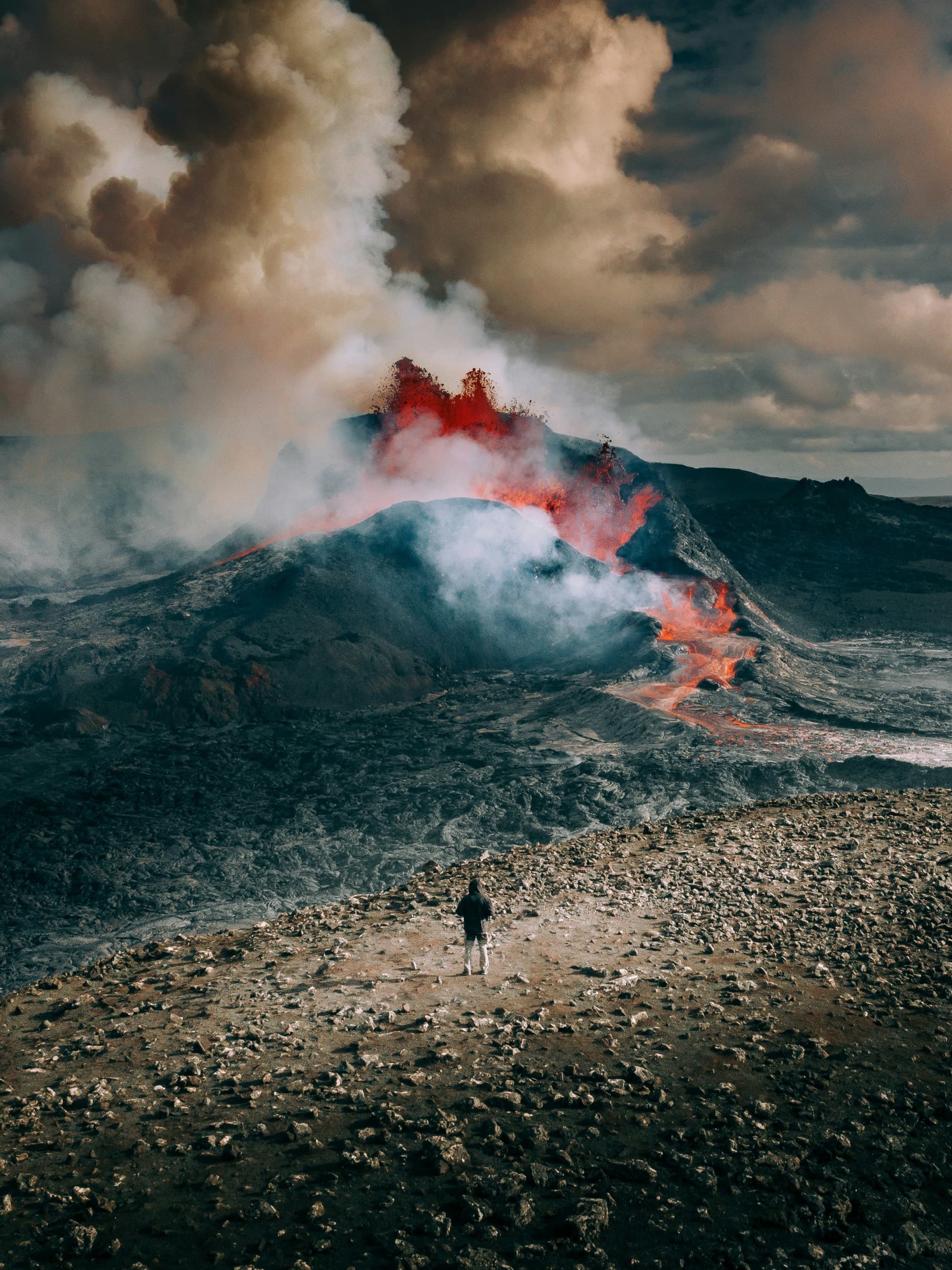 A person standing on rocky terrain with an active volcano and eruptions of smoke in the background.