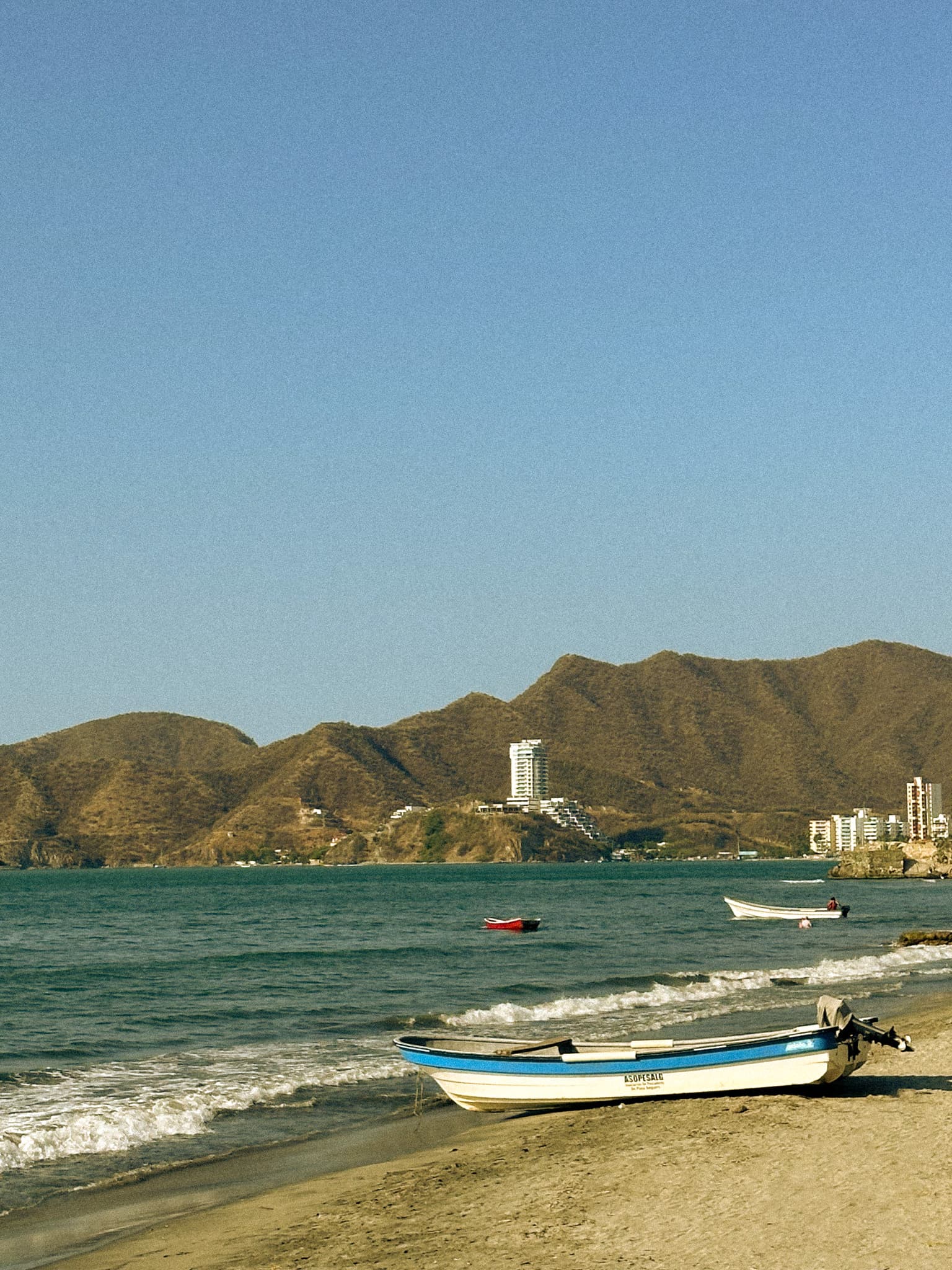 A view of a small boat on a sandy beach shore with the blue water, two boats, city shoreline and brown mountains in the background.