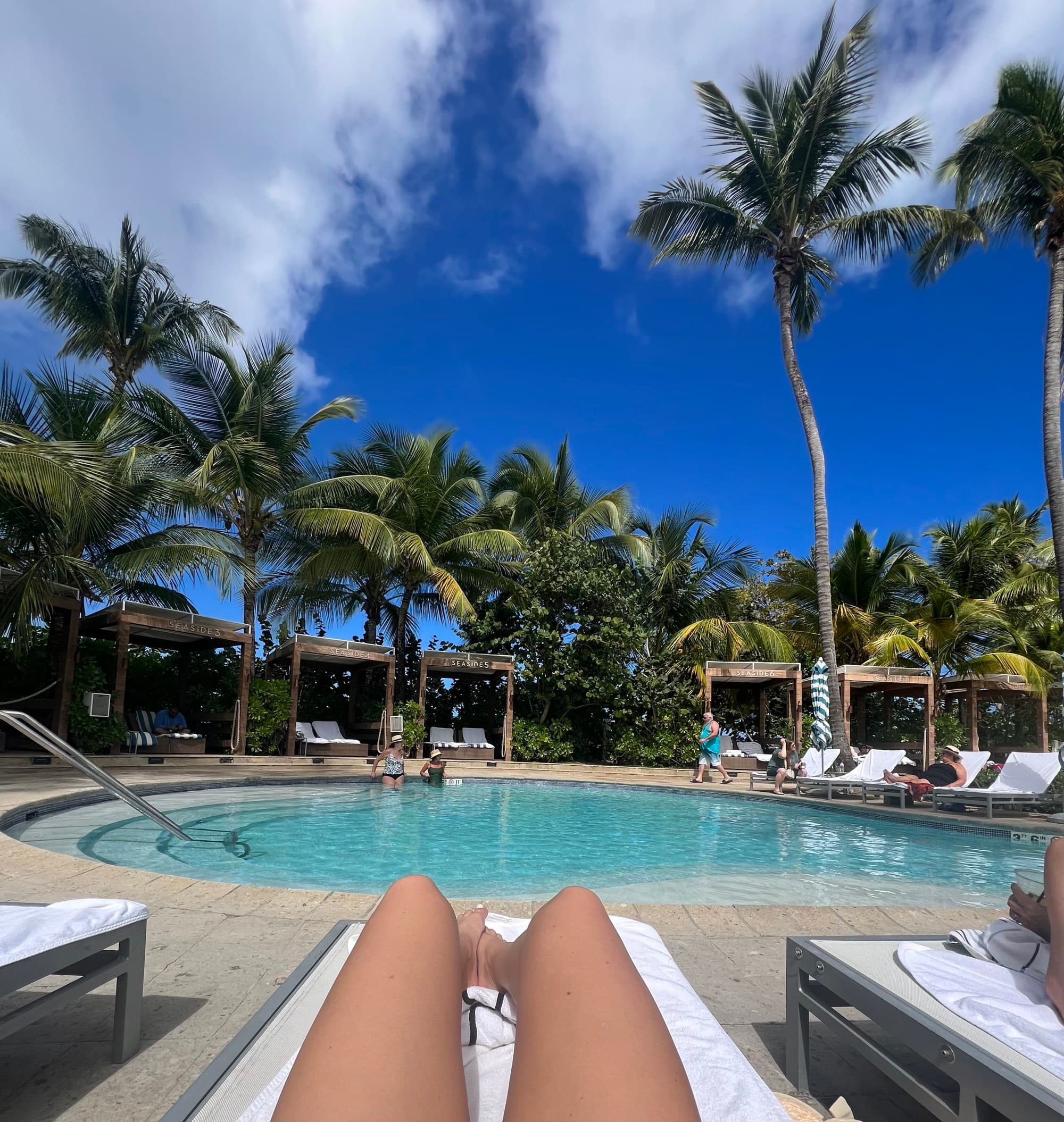 A person tanning their legs on a lawn chair by a swimming pool surrounded by palm trees.
