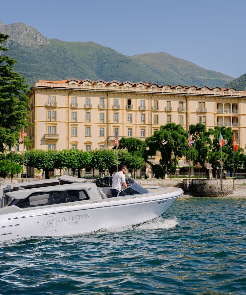 A white boat on Lake Como in front of a luxurious yellow hotel with green trees and mountains.