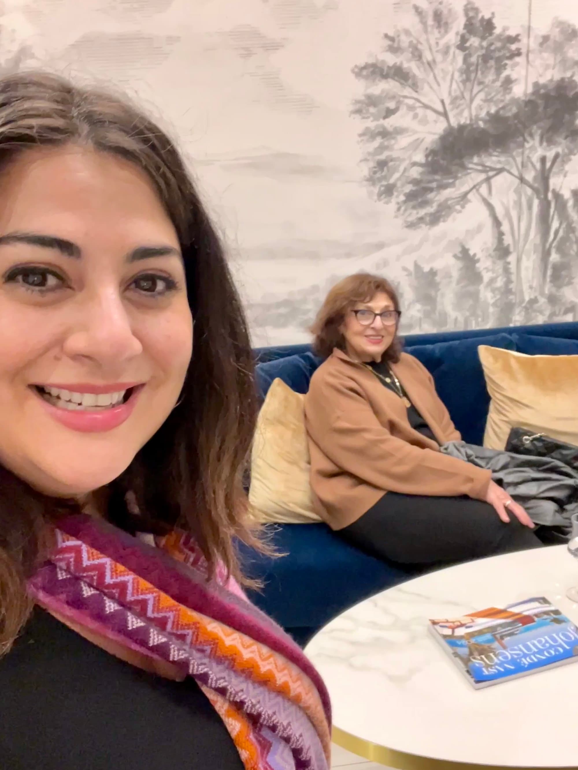 Jennifer posing for a selfie with a person in the background sitting on a blue couch near a white table.
