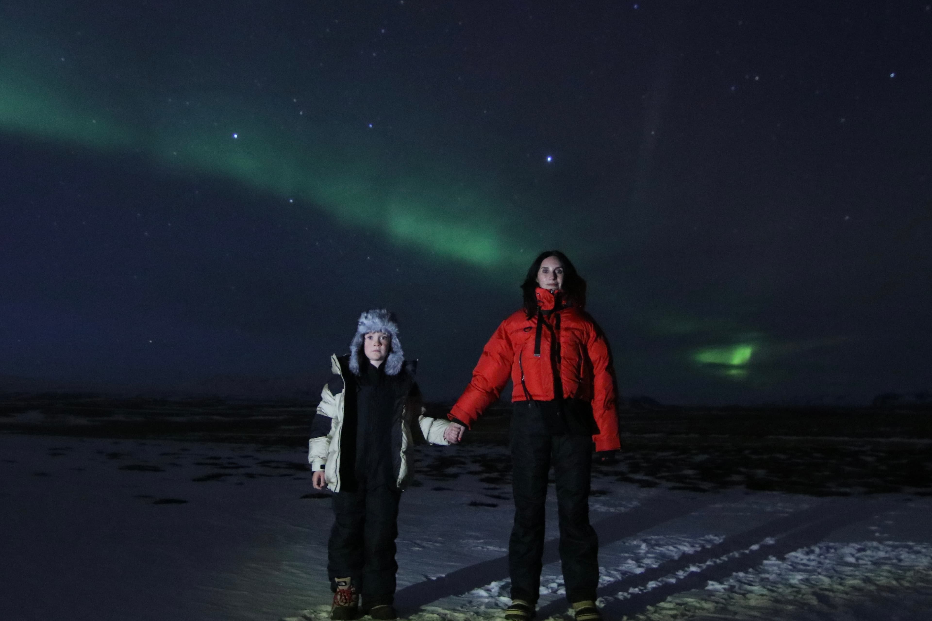 Travel advisor Karen holding the hand of a child, standing on snowy ground at night with the Northern Lights in the sky.